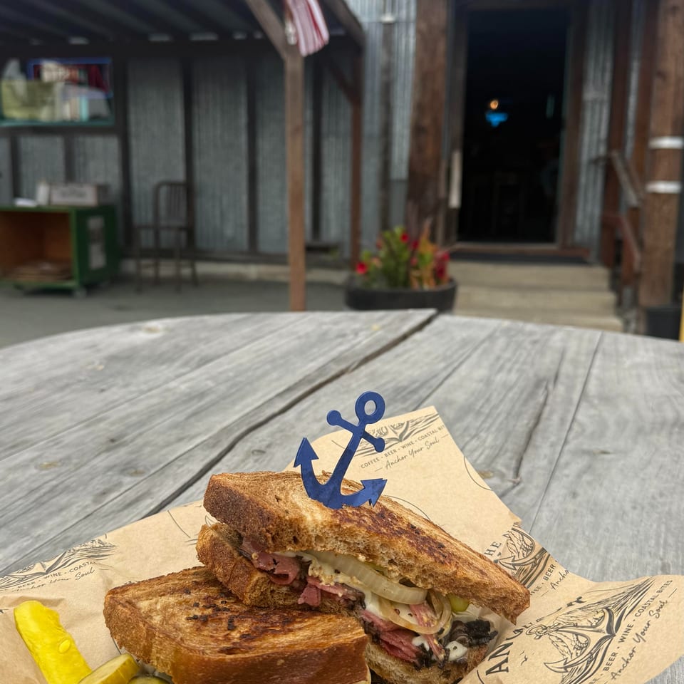 Grilled sandwich with pickles and an anchor pick on a wooden table outside The Nauti Anchor.