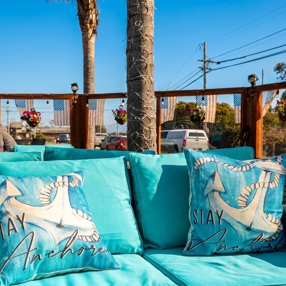 Outdoor patio seating with turquoise cushions and anchor pillows, overlooking a sunny street scene.